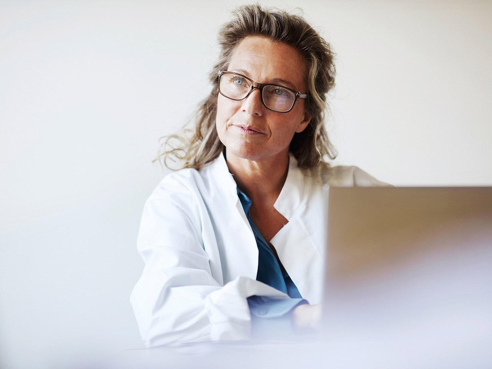A female doctor sits in front of a laptop and looks into the distance